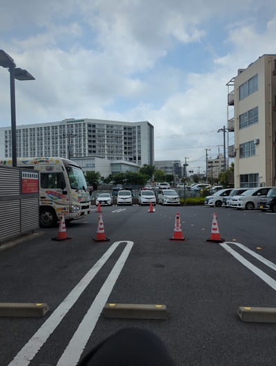 The image depicts a parking lot in Narita, Japan, with a large, multi-story building in the background that appears to be a hospital or medical facility. Several cars are parked in the lot, and a white bus with colorful decorations is parked near the left side of the image. Orange traffic cones are placed on the asphalt, and there are white painted parking lines. The sky is overcast with scattered clouds. There are no people visible in the image.