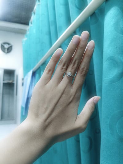 The image shows a close-up of a person's left hand wearing a diamond ring. The hand is positioned in front of a teal-colored curtain. The ring features a central square-shaped diamond surrounded by smaller stones. In the background, a blurry view of a room with shelving and a ventilation fan is visible. The lighting suggests an indoor setting. The location is not specified to be Cileungsi, Indonesia, and no specific details about the environment that would indicate this location are present.