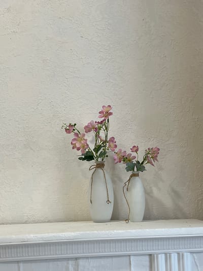 Two white vases with pink flowers stand on a white shelf. The vase on the left is taller and has more flowers. The vase on the right is shorter and has fewer flowers. The flowers are delicate with thin stems and small pink petals. The vases are tied with twine. The background is a textured, light-colored wall. The shelf appears to be part of a fireplace mantel. The scene is indoors, likely in a residential setting in Калодзішчанскі сельскі Савет, Belarus. There are no people or animals present.