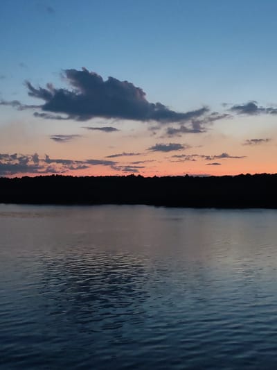 The image depicts a body of water, likely a lake, under a sky transitioning from blue to orange at sunset. A dark treeline sits on the horizon, separating the sky from the water. Clouds are scattered across the sky, with one prominent, darker cloud formation near the top left. The water's surface is rippled, reflecting the colors of the sky and the darkness of the treeline. The location is assumed to be in the vicinity of the city of Даргун, Russia, given the prompt. No people, objects, or actions are apparent other than the environmental scene itself.