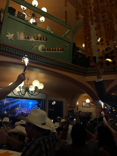 This image depicts an interior scene at the Radio Canta Bar in Guanajuato City, Mexico, during an evening event. The multi-level venue features warm-toned arched architecture and a green-painted balcony bearing the sign "Radio Canta" with "DJ The" text and star embellishments. Numerous strands of orange marigold-like flowers hang from the ceiling as decor. A dense crowd occupies the lower level, with many attendees wearing white cowboy hats. Multiple individuals are holding active sparklers aloft, and several others are recording the scene with smartphones. A large screen in the background displays an event on a stage. A DJ setup, including a laptop, is visible on the upper balcony. Illumination is provided by globe chandeliers, the sparklers, and the screen, creating a festive atmosphere.