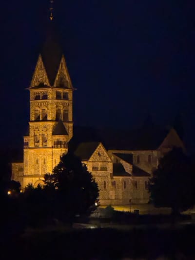 This is a nighttime, eye-level, medium shot of the St. John's Church (Johanniskirche) in Hanau, Germany, illuminated by warm artificial lights against a dark blue sky. The church is constructed of stone and features a prominent, multi-tiered tower topped with a spire. The tower has decorative stonework and multiple arched windows. The main body of the church, with its pitched roof and smaller windows, extends to the right of the tower. Silhouetted trees and foliage are visible in the foreground, obscuring the lower portion of the church and the immediate surroundings. There are no people or discernible animals in the frame. The overall atmosphere is one of quiet solemnity and historical presence.