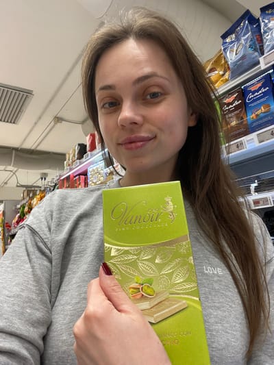 A female individual is depicted in a supermarket in Monsummano Terme, Italy, holding a bright green box of "Vanoir FINE CHOCOLATE" with gold accents. The box front displays illustrations of cocoa pods and two white chocolate bars with pistachios, bearing the partial text "BIANCO CON PISTACHIO". She wears a grey sweatshirt with "LOVE" embroidered on the left chest and has dark red nail polish on her left hand, which holds the chocolate. Her long brown hair frames a face with light-colored eyes, and she exhibits a slight smile directed towards the viewer. The background reveals typical supermarket shelving stocked with various packaged food items, including other chocolate brands like Venchi and Quadro, with visible price tags such as €4.95 and €2.99. Overhead fluorescent lighting and a ceiling ventilation grille are also visible within the store environment.