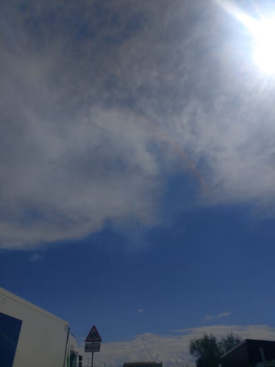The image shows a bright sky with scattered clouds and a faint rainbow arc. The sun is visible in the upper right corner, casting a strong glare. In the lower left, a portion of a white truck is visible. To the right of the truck, a triangular road sign is present, depicting figures of children and an arrow pointing downwards with the text "100m". Further to the right, silhouetted trees and buildings are faintly visible in the distance. The scene appears to be outdoors, possibly on a road, given the presence of the truck and road sign. No specific details about the city of Jrvej, Armenia are discernible from the image itself.