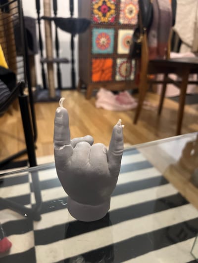 A gray hand-shaped sculpture rests on a glass table with black and white stripes. The sculpture is posed with the index finger and pinky extended, while the other fingers are curled inwards. Remnants of white string or fiber are adhered to the fingertips. In the background, there's a multi-colored, patterned cabinet and wooden furniture.