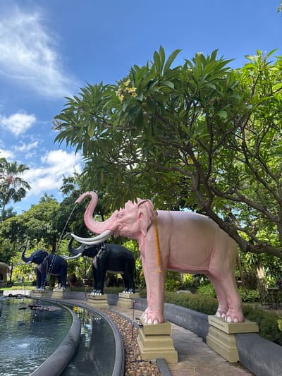 The photograph shows a pink elephant statue as part of a larger display of elephant statues in a park-like setting. The pink elephant is the most prominent element, with a yellow garland around its neck. Black elephant statues are visible behind it near the edge of a multi-tiered water feature. Lush trees with large leaves and a blue sky with scattered clouds are visible in the background. The setting is likely a public space or garden, as suggested by the landscaping and water feature. This is located in the city of Samrong Nuea Subdistrict Municipality, Thailand.