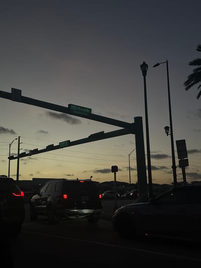 The image displays an urban street intersection at dusk or dawn in Aventura, Florida, United States. The sky transitions from dark grey-blue at the zenith to a lighter orange-gold hue near the horizon, with scattered horizontal cloud formations.
In the foreground, multiple vehicles are stationary on an asphalt road. A dark sport utility vehicle (SUV), likely a Ford Explorer, is positioned centrally, exhibiting illuminated red taillights and bright white light reflecting from its lower rear bumper and quad exhaust tips. To its right, the front portion of a dark sedan is visible. To its left, the rear taillight of another dark vehicle is partially seen.
Overhead, a prominent metal gantry supports multiple signs. A large green sign reads "US 1 / Biscayne Blvd" with "Aventura Pkwy" directly below it. Further to the left on the gantry, a square white sign states "NO TURN ON RED". Below the main gantry, a smaller green sign indicates "NE 199 St".
On the right side of the frame, an ornate, dark street lamp post and a taller, more modern streetlight pole are silhouetted against the sky. Below these, a white rectangular sign reads "DO NOT BLOCK INTERSECTION". Palm trees are discernible on the far right edge of the image. Distant commercial buildings or structures form a dark silhouette against the horizon. No individuals are visible. The scene depicts a typical traffic standstill at an intersection during low light conditions.