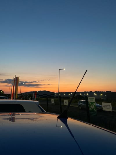This is a vertical, low-angle, outdoor photograph taken at dusk. The scene is primarily a sky transitioning from a deep blue on the left to a vibrant orange and yellow sunset on the right. In the foreground, the polished roof of a dark-colored vehicle reflects the colorful sky. An antenna extends from the roof towards the sky.

In the mid-ground, a chain-link fence runs horizontally across the image. Behind the fence, there are several signs and a building. One sign, in white text on a dark background, reads "KIEROWNIK OCZYSZCZALNI ŚCIEKÓW". Another sign, green with white text and a white symbol of a snowflake with four arrows pointing outwards, reads "OCZYSZCZALNIA ŚCIEKÓW". A third sign, also white text on a dark background, contains Polish text that appears to be "WAŻNY JEST TYLKO NASZ POSTĘP". Streetlights are visible along the fence line, casting a warm glow. German flags, with colors black, red, and yellow, are also visible on poles in front of the fence.

The location is identified as Bolesławiec, Poland. The time of day is clearly dusk, with the sunset dominating the sky. The weather appears to be clear, allowing for a vibrant sunset. The overall mood is tranquil and reflective, enhanced by the beautiful sky and the car's reflection.