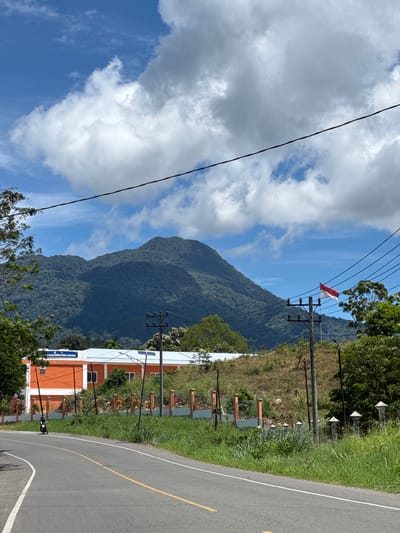 The image shows a scenic view of a road curving towards a lush green mountain under a bright blue sky with scattered white clouds. On the left side of the road, an orange building with white trim, identified by text on its facade as "ISLAMIC BOARDING SCHOOL 27 ACEH BESAR," stands behind a fence. A single motorcycle with a rider is visible on the road in front of the building. A flagpole with the Indonesian flag, red and white, is positioned near a utility pole with wires running across the frame. The landscape is characterized by dense vegetation, including trees and grass, with the mountain dominating the background. The overall scene suggests a tranquil, rural environment in Sukamulia, Indonesia.