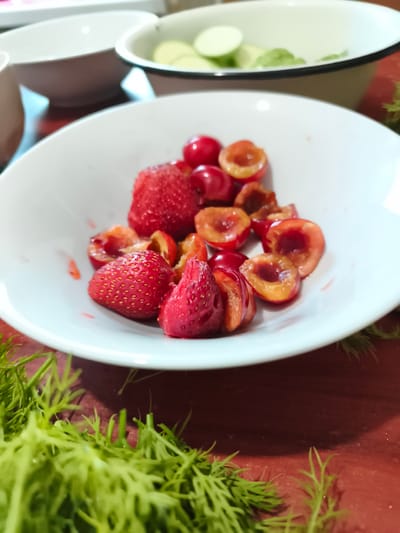 In Sokolivske, Ukraine, a white bowl sits on a dark red wooden surface, filled with sliced strawberries and cherries. The berries are fresh, some whole, some halved, revealing their bright red and orange interiors. Surrounding the bowl, a vibrant green dill garnish is scattered across the table. In the background, another white bowl contains sliced cucumbers. The lighting is soft, highlighting the textures and colors of the fruits.
