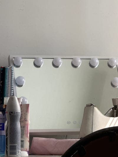 The image depicts a close-up, eye-level view of a vanity mirror with integrated LED lights, positioned in what appears to be a domestic setting, possibly a bedroom or dressing area. The mirror is framed by a white border and is equipped with several round, white LED bulbs. The surface of the mirror reflects various items from the room, including a collection of books and notebooks on the left side, and a light-colored handbag and a pink cloth-like object on the right. In the foreground, several cosmetic products are visible, including a grey aerosol can and a clear bottle with a blue label. The background consists of a plain, light-colored wall, suggesting a minimalist interior. There are no individuals present in the image, and no actions or interactions are depicted. The lighting suggests ambient room light, supplemented by the illuminated bulbs of the mirror. The scene does not contain any elements that would indicate its location in Kovachite, Bulgaria, or any other specific city.