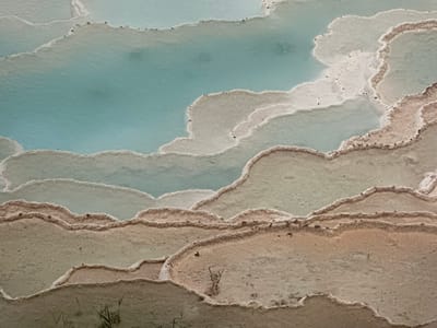 This image presents an abstract, top-down view of travertine terraces, likely a geothermal or mineral deposit feature. The dominant colors are soft blues and greens in the water pooled in the depressions, contrasting with the earthy tones of the surrounding mineral formations. These formations are layered and tiered, with smooth, curved edges that suggest the slow, continuous flow of mineral-rich water. The texture appears granular and slightly porous.
There are no people or discernible objects in the scene, focusing solely on the natural geological formations and water. The lighting suggests daylight, but the time of day and weather conditions are not explicitly indicated. The location context provided is Ossenzijl, Netherlands, which is known for its peat landscapes and wetlands, making this specific geological feature a point of interest.
Notable details include the fine, almost powdery texture of the mineral deposits along the edges of the terraces, and the way the water reflects the sky or surrounding colors. The overall impression is one of serene, natural beauty and geological artistry. There is no visible text in the image.