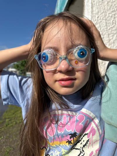 A young girl with long brown hair is lying down outdoors, looking up at the camera. She is wearing novelty glasses with large, cartoonish eyeballs. Her eyes are visible through the lenses of the glasses. She has on a light blue t-shirt with a colorful graphic print on it, featuring pink and purple abstract shapes, palm trees, and the text "SINCE 1993". She is also wearing a light green sweatshirt. The background is blurred, showing a white textured surface, green foliage, and a bright blue sky. The lighting suggests it is daytime with clear weather. There are faint reflections and text visible on the lenses of the glasses, but it is difficult to read. The overall impression is one of playfulness and a casual outdoor setting.