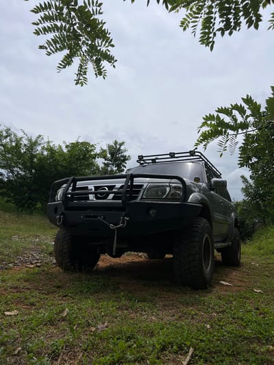 A dark-colored, heavily modified Nissan Patrol SUV is parked on an unpaved, grassy area in Cañacillas Arriba, Panama. The vehicle features large off-road tires, an aftermarket steel front bumper with an integrated bull bar and grille guard, a visible winch with a metal hook/shackle, and a roof rack equipped with four auxiliary LED lights. The front view of the vehicle shows the Nissan emblem on the grille and both headlights. The ground consists of dirt, sparse green grass, and scattered small rocks. Lush green trees and dense foliage line the background, with leafy branches overhanging the top portion of the frame. The sky is overcast with light gray clouds. No individuals are present, and no actions or interactions are depicted.