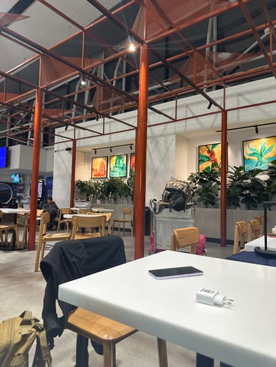 The image shows an indoor space with a modern, industrial aesthetic. An orange steel structure with a geometric pattern dominates the left side of the frame. In the background, there are plants, chairs, and artwork on the walls. At a table, there is a smartphone and a white charging block. A person wearing black is partially visible at the table. Additional tables and chairs are visible in the distance. The overall setting suggests a public space, possibly a café or lounge at an airport, considering the presence of travel-related infrastructure, particularly the location being identified as Pudahuel, Chile. A person wearing black attire is observed sitting at the table.