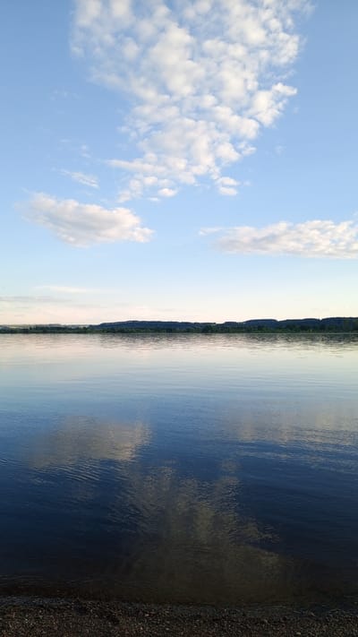 The image displays a tranquil panoramic landscape of a large body of water in Шолья, Russia, under a clear, partially cloudy sky. The upper third of the frame consists of a light blue sky populated by scattered white cumulus clouds, which are denser in the upper left corner and sparser towards the upper right. The horizon, positioned near the middle of the vertical frame, features a distant, dark green landmass characterized by an undulating line of dense tree cover or hills. The lower two-thirds of the image is dominated by a calm expanse of water, likely a river or lake, which clearly reflects the blue sky and white clouds above, albeit with slight distortion from gentle ripples across its surface. The water color transitions from a lighter blue near the horizon to a deeper, darker blue in the foreground. The very bottom edge of the frame reveals a narrow strip of dark, coarse gravel or pebbles along the immediate shoreline. No individuals, boats, or other man-made structures are visible within the scene.