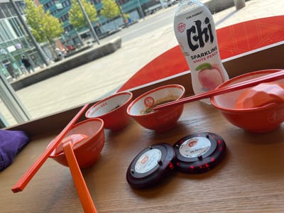 In Bottrop, Germany, a table is set for a meal with several red bowls containing food. Two pairs of red chopsticks rest in the bowls. Next to the bowls are two black wireless restaurant pagers, one displaying red lights. A bottle of "Chi Forest Sparkling White Peach" drink stands upright on the table. The table is positioned in front of a large window, offering a view of a city street with buildings, trees, and people walking by. The overall setting suggests a casual dining experience.