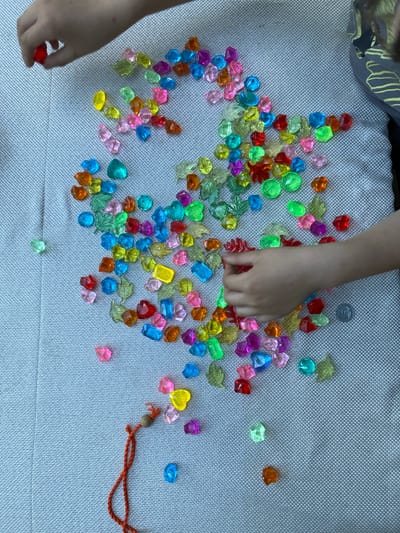 The image shows a collection of colorful, translucent, acrylic gems scattered across a textured grey surface, likely a blanket or outdoor seating cushion. Several of the gems are leaf-shaped. Two children's hands are visible in the frame. One hand, on the left side of the image, is picking up a red gem. Another hand, on the right side of the image, appears to be interacting with the gems. A small, orange string with a wooden bead is also visible in the lower left quadrant of the image. A single coin is positioned near the hand on the right. The scene is lit by natural light, creating shadows on the surface. No specific landmarks or indicators of location in Lobnya, Russia, are present within the image.