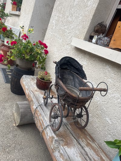 The image depicts a rustic outdoor scene in Les Rousses, France, likely a residential area or garden. The foreground features a weathered wooden beam serving as a bench, with a small, antique-style doll carriage placed upon it. The carriage is made of dark wood with a fabric canopy and metal wheels, secured by a chain. To the left of the bench, a dark wooden barrel is visible, topped with a pot containing red flowers and greenery. Further left, a doormat and more potted plants with red flowers are present. Behind the bench and barrel, a textured, light-colored building wall forms the background. On a windowsill above and to the right of the carriage, several decorative figures are displayed, including two owl figurines and a snail. The overall impression is one of a charming, slightly aged, and decorative outdoor space.
