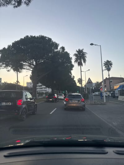 The scene depicts a multi-lane urban street in Cagnes-sur-Mer, France, viewed from within a vehicle at dusk or early evening. The sky transitions from light blue directly overhead to an orange-yellow gradient at the horizon.
In the foreground on the left, a dark grey Smart car (French license plate GL-078-JK) with illuminated taillights is in a traffic lane. Behind it, another dark vehicle and a white car are visible. The left side of the road is bordered by a row of large, dense pine trees and multiple palm trees, which partially obscure white, multi-story residential buildings featuring balconies.
Centered in the lane ahead, a grey Mercedes-Benz (French license plate CZ-187-TP) with illuminated taillights is stationary or moving slowly. The asphalt road surface displays white lane markings.
On the right side of the frame, a red traffic signal is visible at an intersection. A pedestrian wearing a blue top walks on the sidewalk, accompanied by a small, light-colored dog on a leash. A triangular road sign with a white background and black symbol warns of "2 PASSAGES SURELEVÉS SUCCESSIFS" (2 successive raised crossings). Below it, a white A-frame sign displays "7/7" and "7h-18h". Buildings on the right include a two-story structure with reddish-brown walls. A blue and white bus, along with several white vans and other vehicles, are parked or stopped along the right side of the street. Multiple palm trees and active streetlights are also present on the right. Dashboard reflections are visible along the bottom edge of the image.