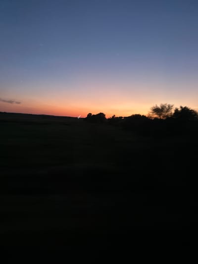 This is a landscape photograph taken at sunset in Zvertiv, Ukraine. The image captures a rural scene with a vast sky transitioning from deep blue at the top to hues of orange and pink near the horizon. A silhouetted treeline runs across the midground, with a large, dark expanse of what appears to be a field or forest dominating the right side of the frame. A faint, white object, possibly a distant airplane or a light streak, is visible against the colorful sky. The overall atmosphere is serene and tranquil, characteristic of the end of the day. The image is oriented vertically.