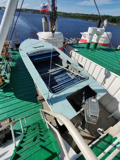 A top-down, slightly angled view shows the deck of a large ship, likely a ferry or cargo vessel, on a sunny day. A smaller, light blue boat with wooden slats is positioned on the green deck, partially obscuring a white structure with red trim. The smaller boat has an outboard motor, a Suzuki 15 horsepower model, mounted at the stern. The deck is painted bright green and shows signs of wear and tear, with some areas appearing darker and possibly wet. White railings and stairs are visible, along with some metallic equipment. In the background, a body of water stretches out, with a forested shoreline and a few buildings visible in the distance. A Russian flag is flying from a mast. The overall impression is of a functional, working vessel on a calm day.