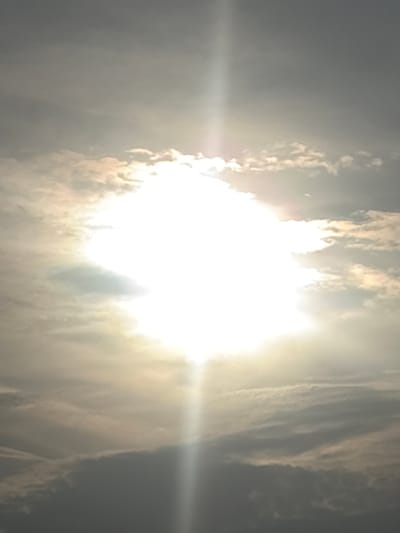 The image captures a bright, sunlit sky, dominated by the intense glare of the sun at its center. The sun appears as an overexposed, brilliant white orb, largely obscuring its shape due to its brightness. Surrounding and partially covering the sun are clouds, which are illuminated from behind or through. These clouds exhibit a range of tones, from glowing golden and bright white where the sunlight penetrates, to darker, muted grays and browns in areas of denser cloud cover.

A prominent horizontal lens flare extends from the left side of the sun across the middle of the frame, indicating direct light entering the camera. The overall impression is of a vibrant and powerful natural light source. The time of day appears to be either midday or late morning/early afternoon, given the high intensity and apparent position of the sun. The weather is partly cloudy, with scattered clouds creating a dynamic interplay of light and shadow in the sky. No people, man-made objects, or text are visible in the frame, making the sky and its celestial elements the sole subjects of the photograph. The image conveys a sense of natural grandeur and the overwhelming power of sunlight.