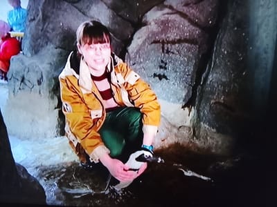 A young person wearing a yellow and white jacket, a striped shirt, and green pants is crouched down in a rocky enclosure. They are holding a penguin with their hands. The person is smiling and looking at the camera. The enclosure appears to be part of an exhibit, possibly at a zoo or aquarium. The background consists of textured rock formations and some water or damp ground. There are also other people visible in the background, blurred and out of focus, some wearing brightly colored clothing.