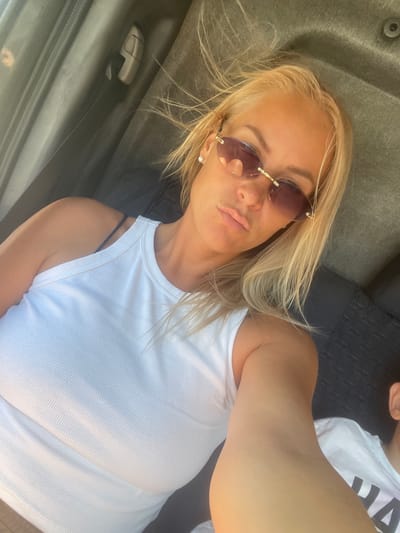 The image shows a close-up of a woman in a white tank top, wearing sunglasses and pearl earrings. Her blonde hair is windswept. She is seated in what appears to be the back seat of a vehicle, with car interior visible in the background. The setting is indoors, within a vehicle. There is no information available in the image to determine the location as Agoy, Russia, or any other specific city.