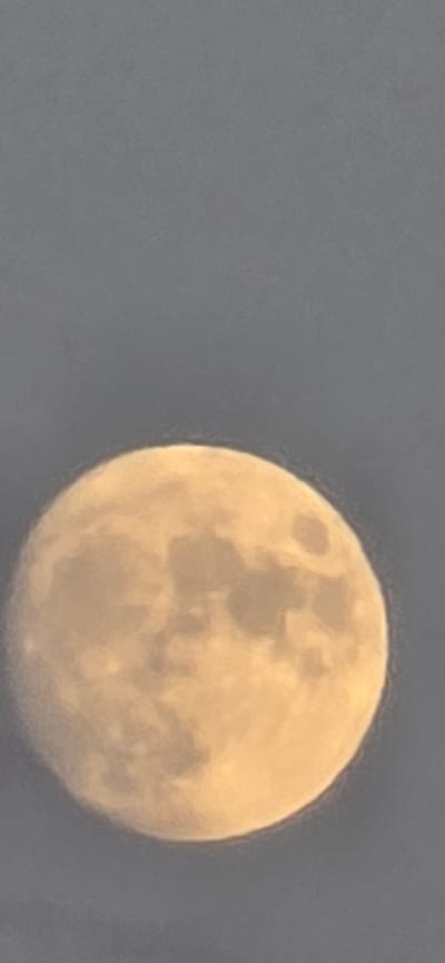 The image shows a full moon against a dark gray sky. The moon is a pale yellow-orange and appears slightly out of focus, with visible surface features that suggest craters and mare. The sky is a uniform shade of gray with some subtle variations in tone, hinting at atmospheric conditions. No specific location in Mazyr, Belarus, can be identified from this image, as it focuses solely on the celestial body.