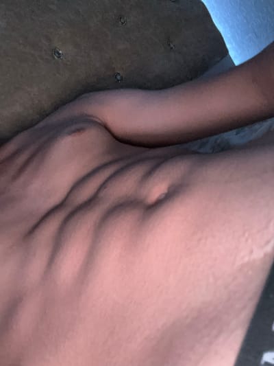 The image shows a close-up view of a person's abdomen, highlighting their well-defined abdominal muscles. The person is lying down, possibly on a textured surface that appears to be a couch or bed. The lighting is dim, casting shadows that accentuate the musculature. The color palette is warm, with tones of pink and beige dominating the skin. The background is out of focus and appears to be a wall with a blue-tinted light source. There is no indication of the location in Ognianovo, Bulgaria, or any specific context beyond the subject's body.