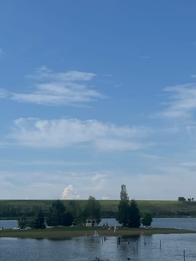 The image depicts a wide shot of a body of water, likely a lake, under a bright blue sky with scattered white clouds. The scene is oriented vertically, suggesting it was taken with the camera held sideways.
The water's surface is calm, reflecting the sky and surrounding landscape. Along the edge of the water, there is a strip of green grass and trees. In the distance, a vast, green, open field stretches towards the horizon. Several small figures, appearing to be people, are visible along the grassy bank and in the water, some engaged in activities. There are also some structures, possibly docks or small buildings, near the water's edge.
The lighting suggests it is daytime, likely mid-morning or afternoon, given the bright sky. The presence of clouds indicates a partly cloudy day. The location is identified as Emelyanovskoe Lake in Russia. No text is visible in the image. The overall impression is one of a recreational outdoor setting, possibly a day at the lake with people enjoying various activities.