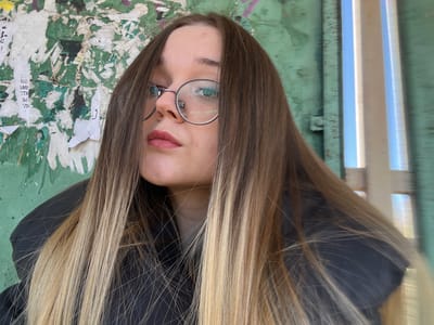 An image depicts a close-up, low-angle portrait of a young adult female in the city of Большевик, Russia. The subject has long, straight, ombre hair, transitioning from medium brown to blonde, framing her face and resting over her shoulders. She wears round, silver-rimmed glasses, with reflections visible on the lenses. A silver circular nose ring is present on her left nostril. Her lips are colored with pink lipstick, and she looks directly at the camera with a neutral expression. She is partially visible, wearing a dark-colored jacket or coat.
The background to her left features a distressed wall with peeling green paint, heavily covered by multiple layers of torn white paper posters, some showing faint Cyrillic text. To her right, the background transitions to a smoother, muted green surface, followed by several vertical, reflective metallic structures that appear motion-blurred, showing reflections of a bright blue sky and white elements. The lighting suggests natural daylight.