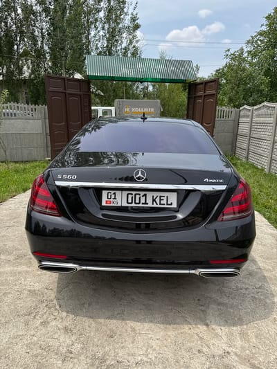 In КРУГОВО, Kyrgyzstan, the rear of a black Mercedes-Benz S560 4MATIC is parked on a concrete surface. The car features tinted windows, distinctive taillights, and dual exhaust pipes. A license plate with the numbers "01 001 KEL" and the abbreviation "KG" is visible. Behind the car, a brown wooden gate stands open, leading to a corrugated metal structure. Concrete fences and lush green trees form the surrounding environment under a cloudy sky. A partial view of a sign with "HK KOLLMER" is also present.