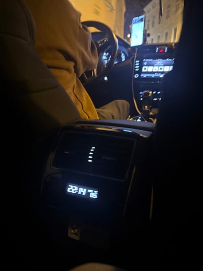 The image captures the interior of a vehicle at night, observed from a rear passenger perspective, within the city of Krems an der Donau, Austria. In the foreground, the rear climate control unit is prominently visible, displaying the time "22:14" and a temperature of "8.5°," accompanied by five illuminated vertical indicators, likely representing fan speed. A small yellow triangular symbol is present below this display.
A person, identified as the driver, occupies the front left seat, wearing a light brown or tan hooded garment and dark trousers. Their hands are positioned on or near the steering wheel. A smartphone is mounted on the dashboard, displaying a map or navigation application. The vehicle's dashboard and center console are illuminated, revealing a digital infotainment system with various buttons and controls, and a visible gear selector area. Through the front windshield, blurred, illuminated building facades are discernible, indicating an urban setting at night. The scene is primarily lit by internal vehicle illumination and external street lighting.