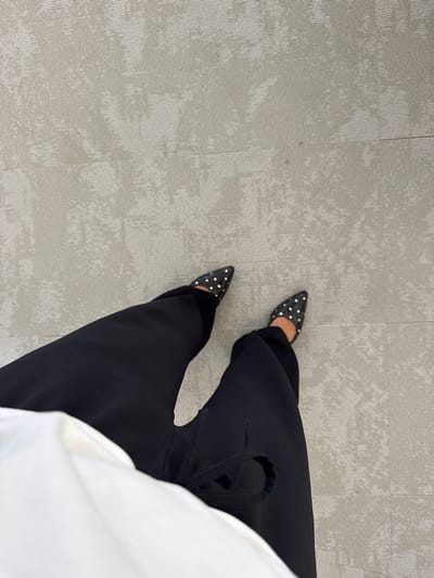 The image shows a top-down view of a person's legs and feet wearing black pointed-toe flats adorned with white polka dots. The individual is dressed in black wide-leg pants and a white top. The shoes are positioned on a light gray carpeted surface, which appears to be the floor of a room. There is no specific indication of the location being in the city of Les Arcs, France; the scene is generic and could be anywhere with this type of flooring. The lighting is neutral and even, suggesting an indoor environment.