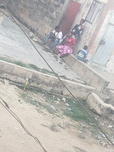 A ground-level, slightly out-of-focus shot shows a group of people sitting outside a building in Alalubosa, Nigeria. A man in a white shirt and jeans is seated on a step. Next to him is a person in a red shirt, followed by another in a pink patterned dress. Further along, a woman in a black floral top stands. Lastly, there's a person in a blue striped shirt seated next to the building. The building itself has boarded-up windows, a red door, and aged brickwork. Adjacent to the building is a patch of dirt with sparse grass and litter. Two power lines run across the foreground. A yellow bird is near the curb.