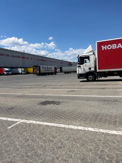 The image shows a wide, paved area in front of a large industrial building in Nerubais'ke, Ukraine. The building, which appears to be a warehouse or distribution center, has a gray facade with red window frames and numerous loading docks. Several trucks are parked in the paved area, some with their trailers open, indicating active operations. One prominent truck in the foreground is white with a large red trailer featuring white lettering that reads "HOBA". Another truck has the text "Peter Rinkleib TRANSPORT GMBH" on its side. The ground is paved with interlocking hexagonal concrete blocks, and white lines mark parking or driving lanes. The sky is clear and blue with scattered white clouds, suggesting a sunny day. The overall scene depicts a logistics or transportation hub.