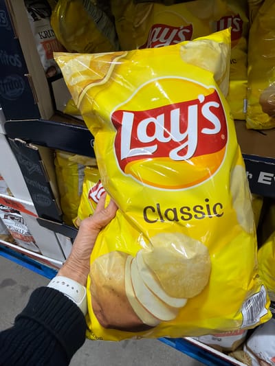 A close-up, eye-level shot shows a person's hand holding a large, yellow bag of Lay's Classic potato chips. The bag has the Lay's logo prominently displayed in red and white. To the left of the bag, other chip bags, including Doritos, are visible on a shelf. The background shows more chip bags on shelving units, suggesting a retail environment such as a supermarket or warehouse store. The overall lighting is bright and even, characteristic of indoor retail lighting. The location is confirmed as Laguna Niguel, United States of America, though the specific store is not identified.