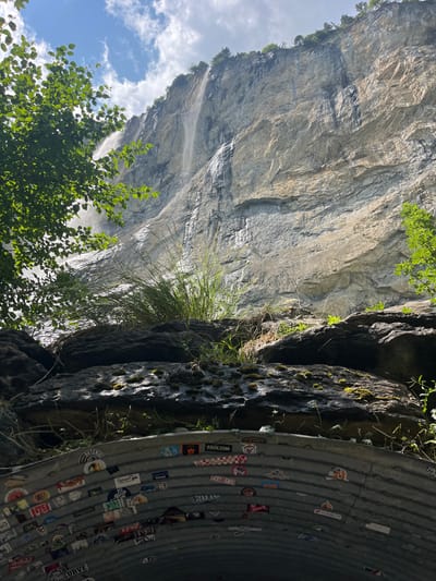The image shows a view looking up towards a sheer, layered rock face with a thin waterfall cascading down its side. Lush green foliage frames the left side of the scene, and other green plants and moss grow from the rocky outcrops in the foreground. Below the rocky overhang, a curved, corrugated metal structure, likely part of a tunnel or drainage system, is visible. This structure is extensively covered in colorful stickers. The sky above is blue with scattered white clouds, and sunlight illuminates parts of the rock face. This view is characteristic of the area near the Staubbach Falls in Wengen, Switzerland, a popular tourist destination known for its dramatic waterfalls and alpine scenery. There are no people or animals visible in the image.