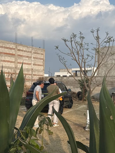 The image portrays an outdoor scene in Sanctorum, Mexico. The sky features large white cumulus clouds on the left and a darker, overcast area on the right. The foreground is dominated by several large agave plants. In the midground, a black SUV is parked on dry, light-colored earth. Three individuals are clustered around the left-rear of the vehicle. A male with grey hair, wearing a light-colored polo shirt and light trousers, stands on the left, oriented towards the other two. An individual with dark hair, wearing a dark top and light trousers, stands with their back mostly to the camera, near the vehicle. A third person, partially obscured, is visible in a light top, holding a yellow-orange object. The SUV's rear license plate displays "07-8." The background includes an unfinished multi-story concrete block building with exposed rebar on the left, a sparsely leaved tree to the right, a low concrete wall, and distant residential structures. A discarded tire lies near the tree.