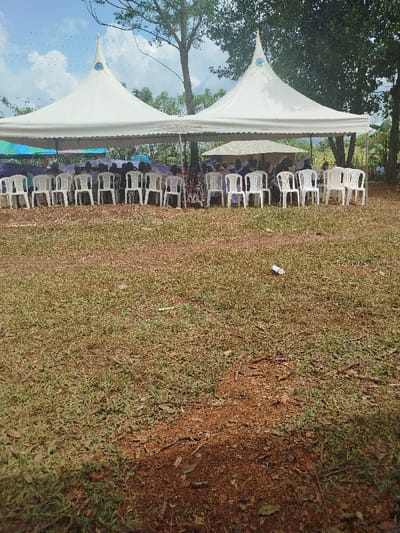 The scene is an outdoor gathering located in North Ugenya ward, Kenya. Two prominent white pyramid-roofed canopy tents are erected over an area of dry grass and reddish-brown soil. Beneath these main tents, multiple rows of white plastic chairs are arranged. Numerous individuals are seated in these chairs, partially obscured by shadows; some are holding light-colored and blue umbrellas. A smaller, lighter-colored tent is visible behind the primary canopies. The ground in the foreground consists of sparse grass, exposed soil, scattered leaves, and small branches. A white plastic bottle lies discarded on the ground in the mid-foreground. Large trees with green foliage define the background, under a partly cloudy blue sky.