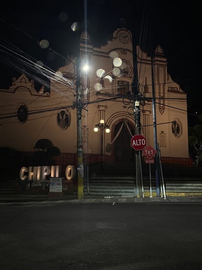 The image shows a vertical view of a street scene in Chipilo, Mexico, at night. A church with ornate white and brown trim dominates the left side of the image. An "Alto" (Stop) sign and a "1X1" sign are visible on a pole in front of the church. Numerous electrical wires are strung across the foreground. On the right, a sidewalk and street are visible. The word "CHIPILO" is spelled out in large, illuminated block letters on the sidewalk.