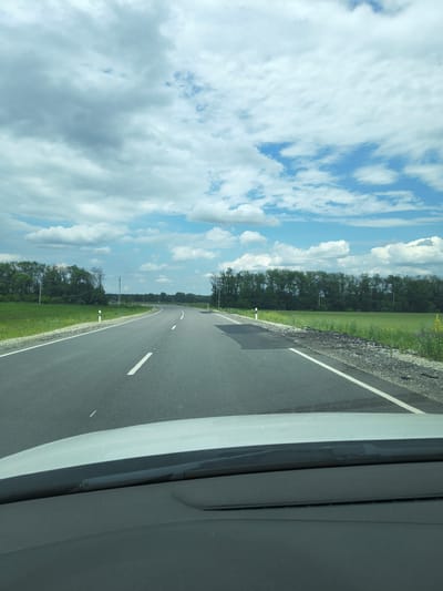 The image shows a view from inside a vehicle, looking out at a two-lane asphalt road curving to the left. The road has white dashed lines marking the lanes. On either side of the road are grassy fields and a line of trees. The sky is filled with fluffy white clouds against a bright blue background. A portion of the vehicle's dashboard is visible in the foreground. The scene appears to be in a rural area, possibly near Zol'skaya, Russia, given the presence of agricultural fields and the type of road. There are no people or animals visible in the image. The weather appears to be clear and sunny with scattered clouds.