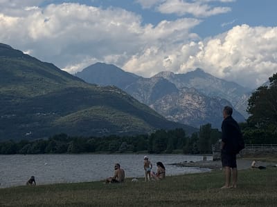 This is a medium-shot, outdoor, daytime photograph taken from a grassy area overlooking a body of water. In the foreground, a man wearing a black short-sleeved shirt and dark shorts stands with his back to the camera, looking out towards the water. His hands are in his pockets. To the left and slightly in front of him, a family of three is seated on the grass. A man with sunglasses sits on the left, a young child wearing a white cap stands in the middle, and a woman in a bikini sits on the right. The water in the middle ground is choppy, with several people visible in and near it. A person in dark swimwear is partially submerged in the water on the left side of the frame. Further to the right, closer to the shore, two people are standing near the water's edge. The background features a dense line of dark green trees, beyond which are layers of imposing mountains. The mountains are dark and rugged, with lighter, rocky peaks visible in the distance. The sky above is a mix of blue sky and dramatic white and grey clouds. The scene appears to be a lakeside recreational area in Colico, Italy, with the prominent mountains likely being the Alps.