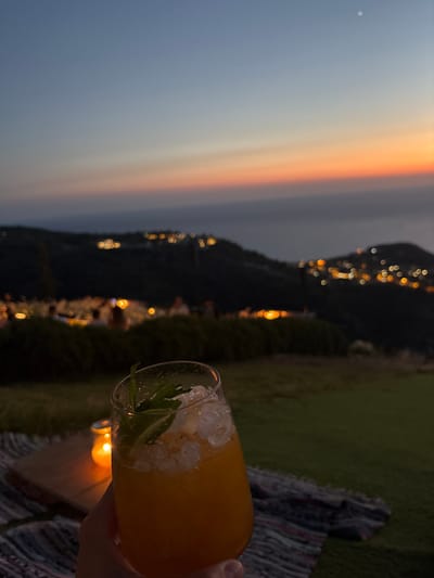 A hand holds a clear glass filled with an orange beverage and ice, garnished with mint, with a blurred background of a dusky sky and distant city lights on rolling hills. The scene appears to be set outdoors at dusk, possibly on a picnic blanket, with a lit candle visible on a wooden surface. The horizon shows a gradient of blues and oranges, transitioning into the dark silhouettes of hills dotted with illuminated structures. The Municipal Unit of Sfakiates, Greece, is known for its mountainous terrain and traditional villages, which aligns with the visual elements of the image.