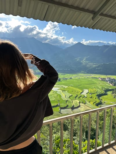 The image depicts a young woman, viewed from behind, standing on a balcony or elevated platform, gazing out at a mountainous landscape with terraced rice paddies. The scene is located in Tú Lệ, Vietnam.

In the foreground, a corrugated metal roof extends across the top of the frame, partially obscuring the sky. Below this, a silver-colored metal railing runs horizontally across the lower mid-section of the image, defining the viewing area. The woman, with long, wavy brown hair, is silhouetted against the bright landscape. She wears a dark grey or black long-sleeved top, possibly a hoodie, and dark pants. Her right arm is raised, with her hand near her forehead, possibly shielding her eyes from the sun or adjusting her hair. Sunlight catches the edges of her hair and the fabric of her sleeve, creating a halo effect.

The midground reveals an expansive valley filled with vibrant green rice terraces, some reflecting water. A winding river or stream traverses the valley floor. Several buildings, indicative of a small town or village, are visible nestled amongst the fields and at the base of the mountains.

In the background, multiple layers of verdant mountains rise under a bright blue sky. Fluffy white clouds are scattered across the sky, with visible sun rays piercing through, creating patches of light and shadow on the distant slopes. The overall impression is a scenic, sunlit view over a rural Vietnamese landscape.