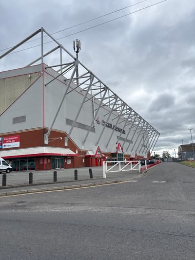 This is a full-shot, eye-level, outdoor, daytime photograph of Mornflake Stadium, located in Crewe, United Kingdom. The stadium, also known as Gresty Road, is a football stadium with a predominantly red and white exterior. A portion of the grandstand, featuring a metal support structure and grey cladding, dominates the left side of the frame. Red trim lines the upper edge of the building and the roofline.
In front of the stadium, a paved area serves as a parking lot. A white van is parked on the left, and several cars are lined up further down the road on the right. A white barrier gate stands across the entrance to the parking area. Black bollards line the edge of the paved area closest to the stadium. The sky is overcast and grey, with a few scattered birds visible. A cell tower is mounted on the stadium's roof. A banner on the left side of the building reads "Proud supporters of The Alex."