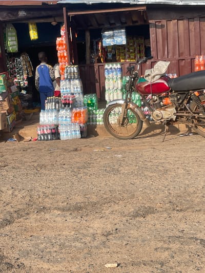 The image depicts an outdoor market stall in KOMU, Nigeria, likely during the day. A man wearing a blue jacket and a beige shirt is standing in front of the stall, which is overflowing with various bottled beverages and packaged goods. Stacks of water bottles, some clear and some with green labels, are prominently displayed. There are also cases of orange drinks and dark-colored sodas. Behind the man and the beverages, a motorcycle is parked, leaning against the stall. The stall itself appears to be constructed from wooden planks. The ground is a mixture of dirt and gravel, suggesting an unpaved area common in outdoor markets. The scene is lit by natural sunlight, casting shadows, indicating daytime. No specific weather conditions are discernible, but the lighting suggests a sunny day. The overall impression is one of a bustling marketplace where goods are being sold.