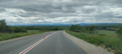 The image shows a grey asphalt road winding through a rural landscape near Radomirtsi, Bulgaria. The road has a wide white line on the left side and orange and white striped cylindrical poles. Lush green fields and low hills stretch out on both sides of the road, receding into distant blue mountains under a heavy, overcast sky. Green vegetation flanks the road, blending into the surrounding scenery.